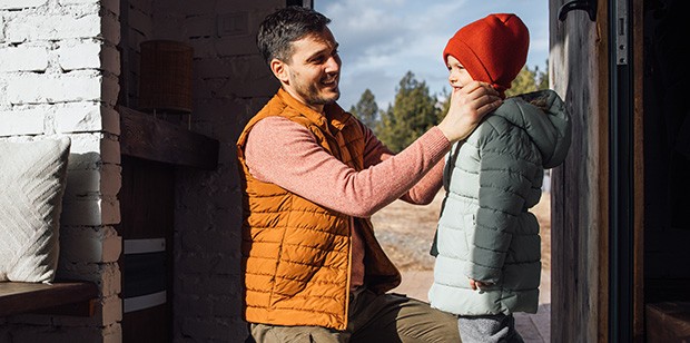 A father helps his child put on a winter coat. Warm sunlight streams through the window, highlighting their joyful expressions.